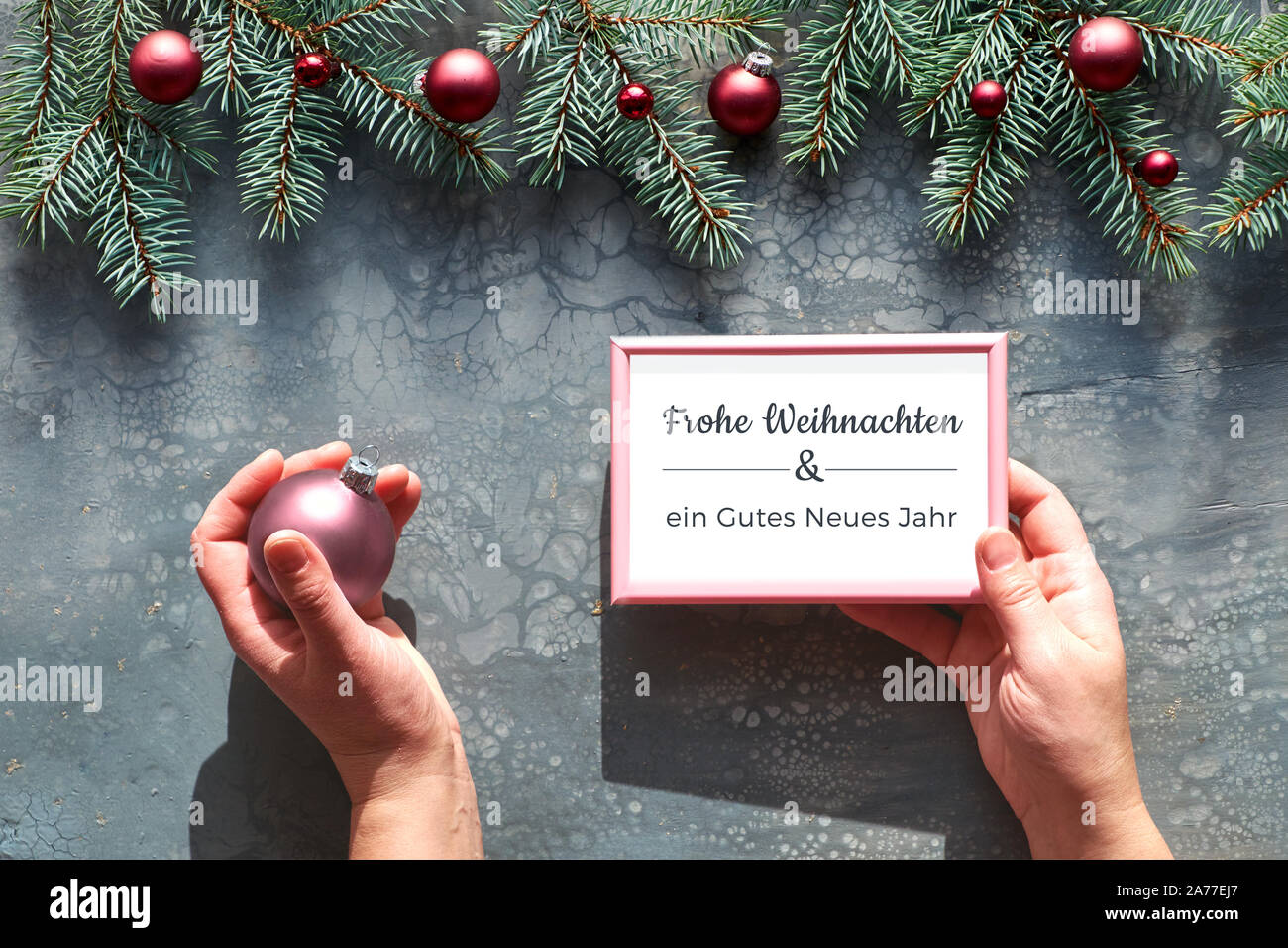 Creative Christmas flat lay on grey acrylic fluid art board. Hands holding pink trinket and frame with text in German "Merry Christmas and a Happy New Stock Photo