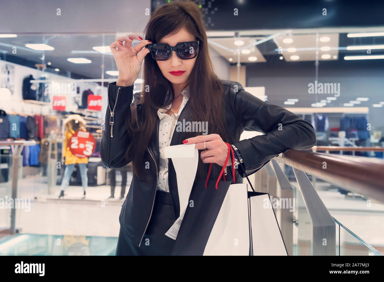 Fashionable woman in dark glasses having good look at long paper with printed bill at shopping background. Shopaholic expenses. Girl with red lips che Stock Photo