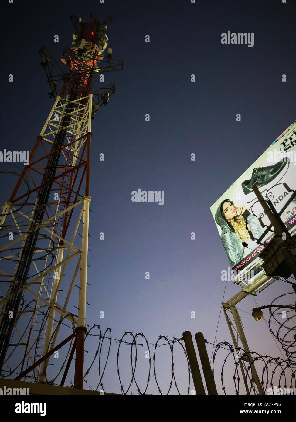 A radio control tower, twisted barb wire fence and a billboard shoe advertisement in Leon, Guanajuato, Mexico at night time. Stock Photo