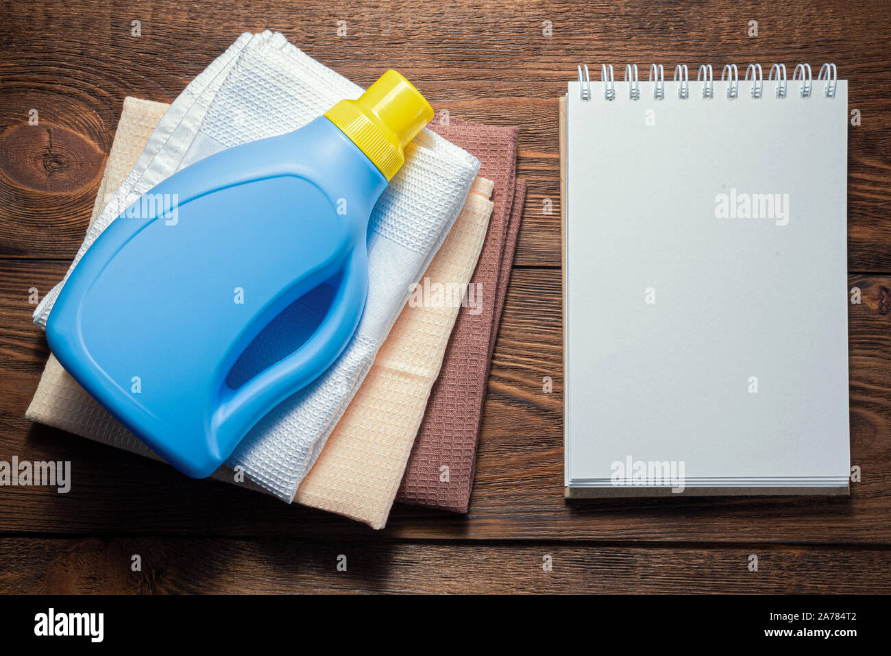 Washing supply, softener, detergent, dish cloth and blank page notepad on wooden table. Household tips mock up. Stock Photo