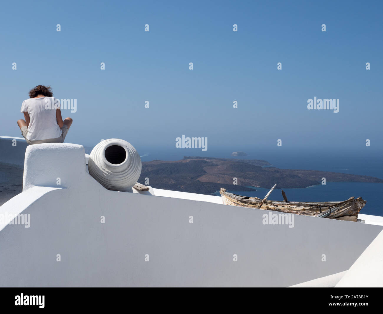a view of woman sitting wall lwith vase pot salvaged, repurposed rowing boat on top a white wall distant mountains horizon sea blue sky Fira Santorini Stock Photo