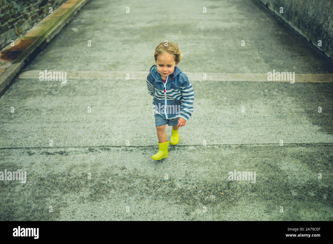 A little toddler in weatherproofs is running on a concrete path Stock