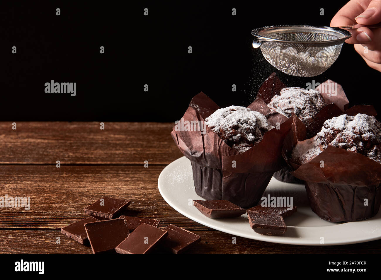 Still life on old wooden board table with three cocoa muffins with sugar and pieces of chocolate. Female hand holding strainer on black background. Sp Stock Photo