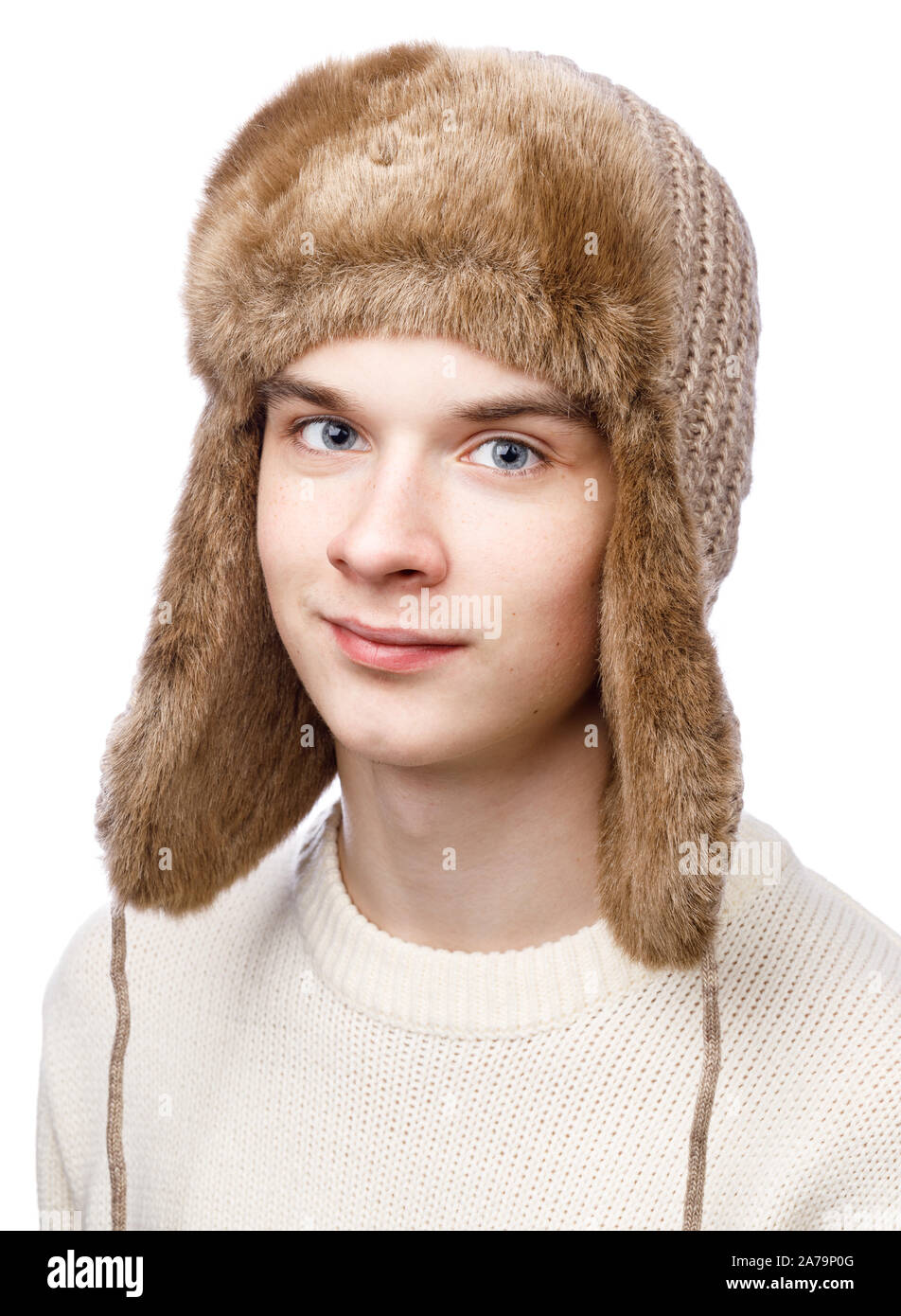 Head and shoulders studio portrait of smiling teenager boy wearing brown knitted faux fur winter trapper hat (Ushanka) and sweater on white background Stock Photo