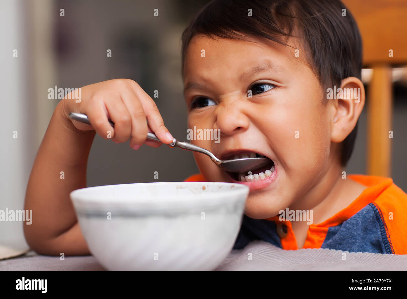 A latino boy taking a bite out of a spoonful of food and sitting unsupported at the dinner table for breakfast. Stock Photo