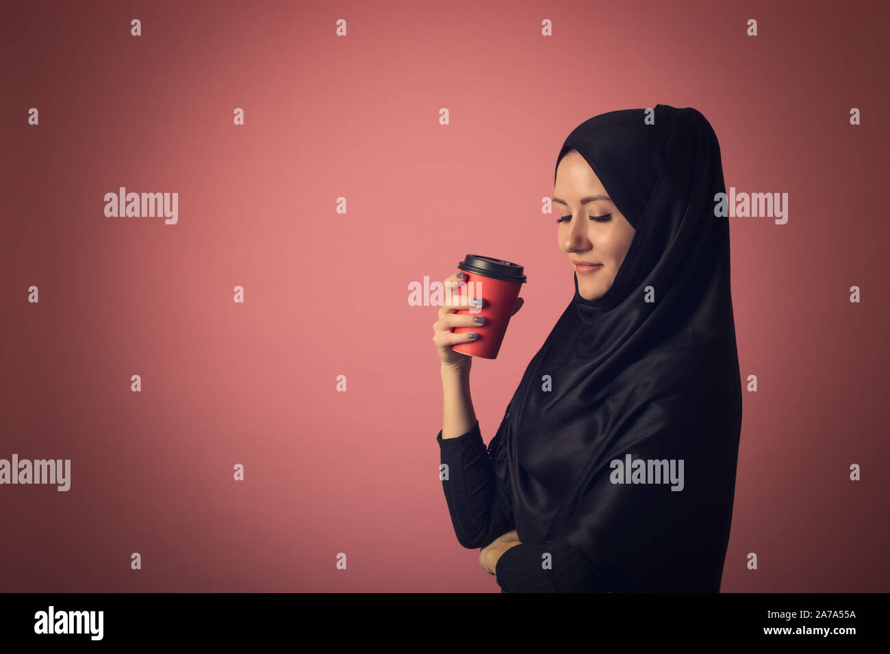 A beautiful Muslim woman with a mysterious look is drinking coffee from a red cup on a pink background in the studio. Concept for street advertising o Stock Photo