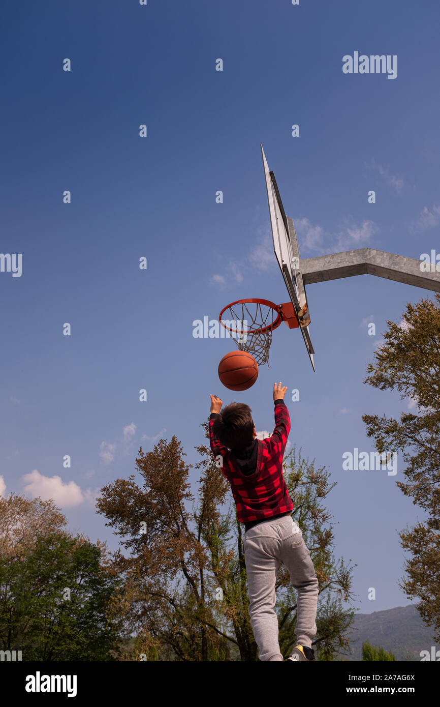 Rear view of a 8-9 years old boy, mid air, shooting jump shot on basketball court. The child has brown hair and is clothing a red sweatshirt. Stock Photo