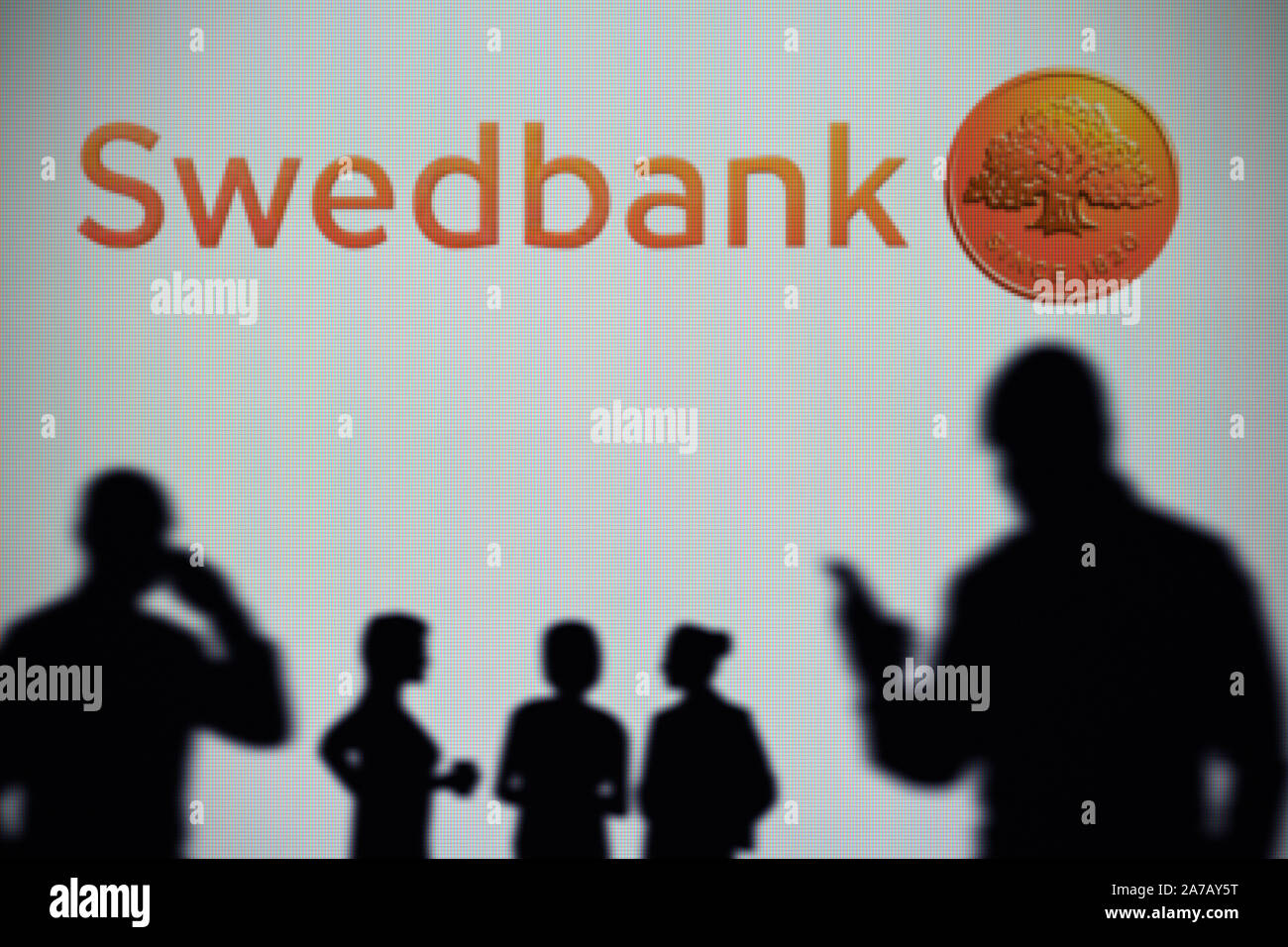 The Swedbank logo is seen on an LED screen in the background while a silhouetted person uses a smartphone (Editorial use only) Stock Photo