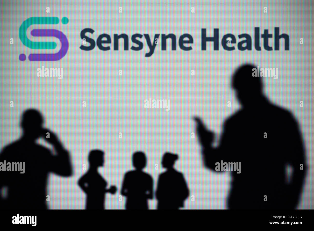 The Sensyne Health logo is seen on an LED screen in the background while a silhouetted person uses a smartphone (Editorial use only) Stock Photo