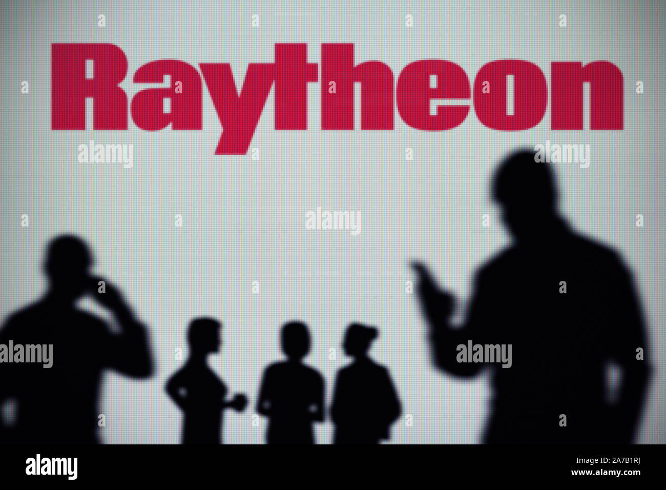 The Raytheon logo is seen on an LED screen in the background while a silhouetted person uses a smartphone (Editorial use only) Stock Photo