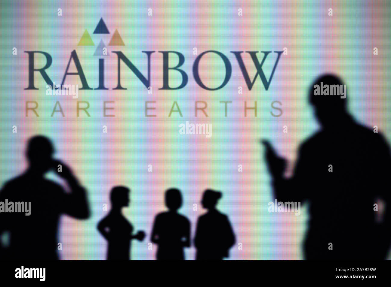 The Rainbow Rare Earths logo is seen on an LED screen in the background while a silhouetted person uses a smartphone (Editorial use only) Stock Photo