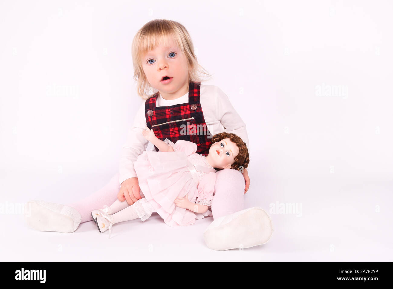 Little blonde toddler girl with big Blue Eyes in red dress play with old doll. Sitting on the floor, white background. Portrait, close-up isolated Stock Photo