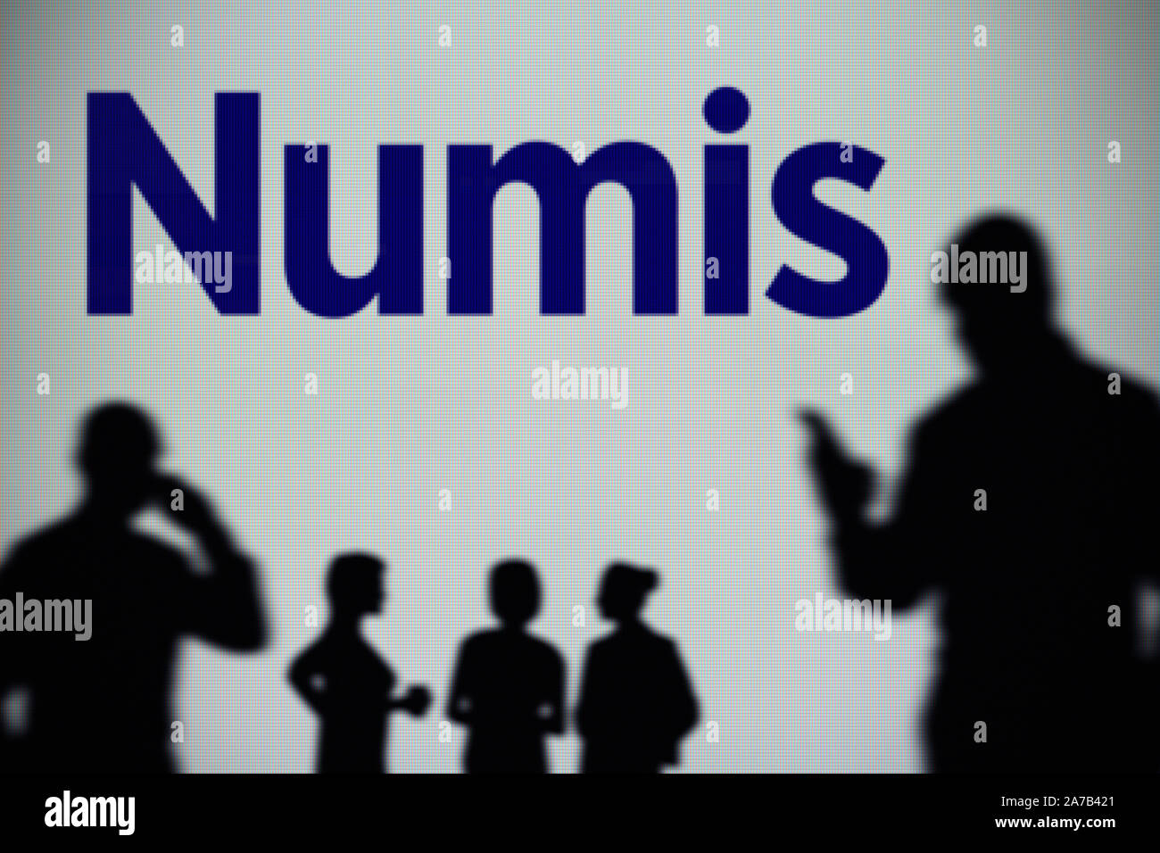 The Numis Securities logo is seen on an LED screen in the background while a silhouetted person uses a smartphone (Editorial use only) Stock Photo