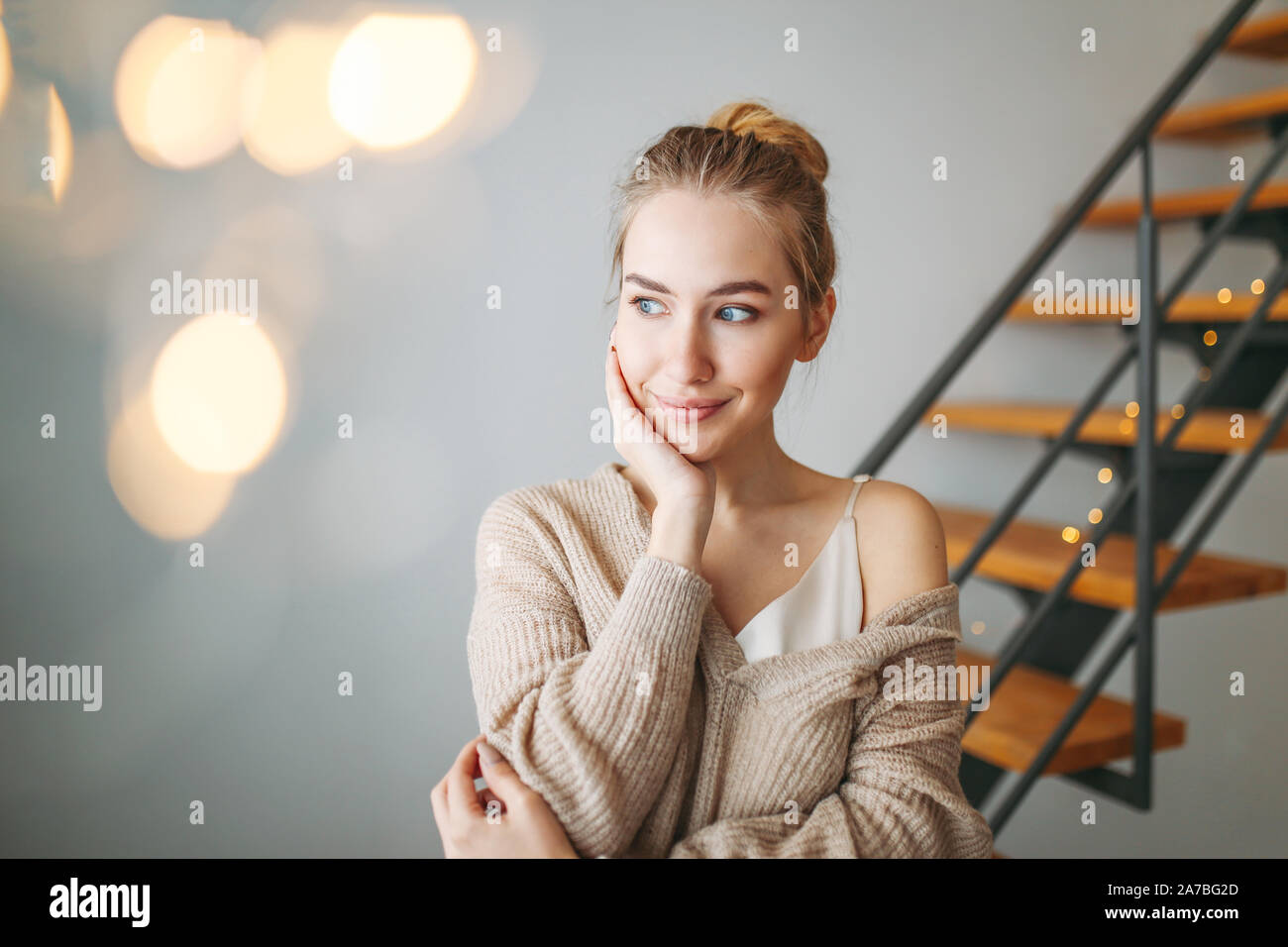 A young beautiful thoughtful girl with blonde hair in an evening silk dress and a cozy cardigan is dreaming about something at home against the backgr Stock Photo