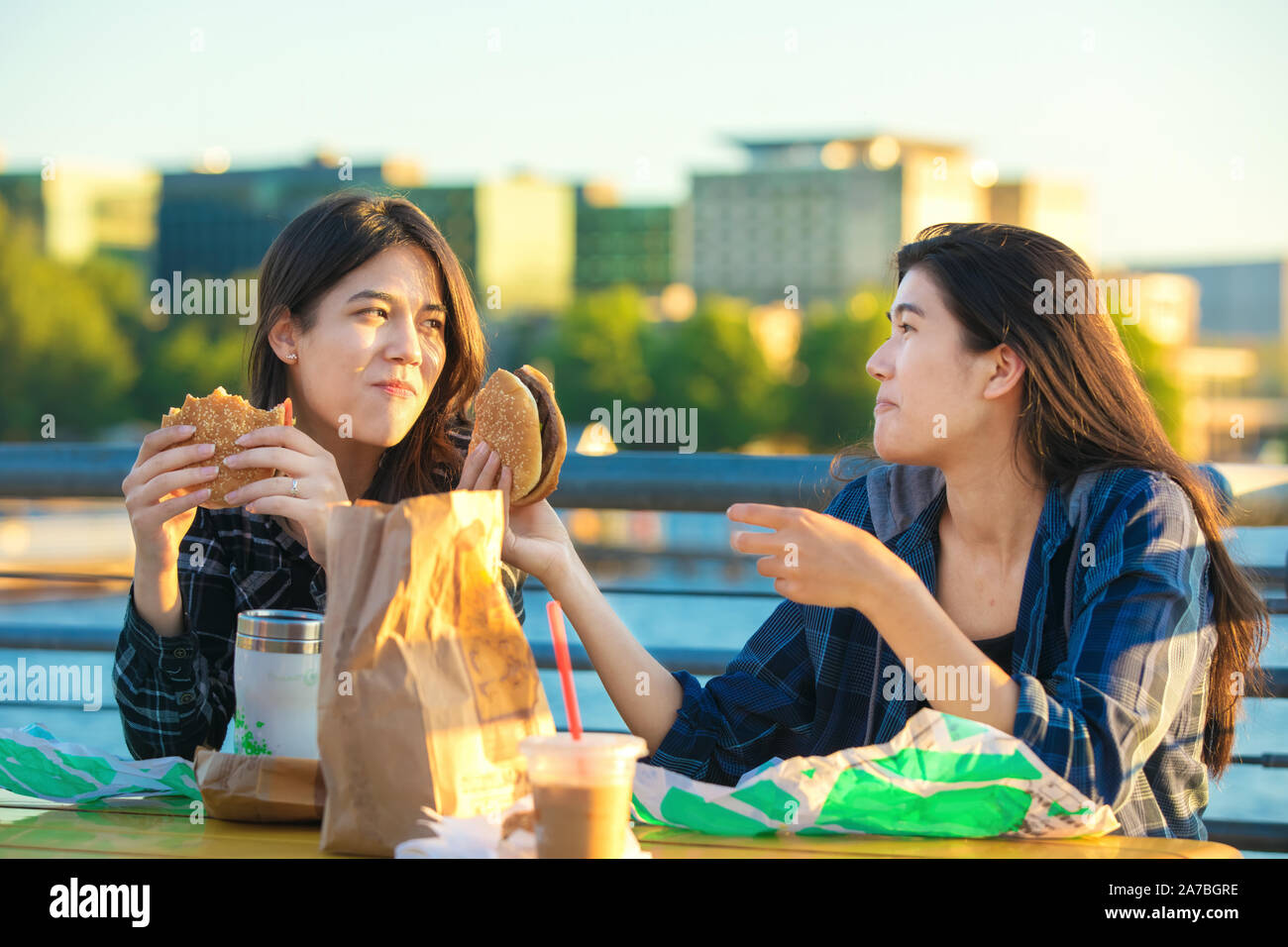 Two biracial Asian, Caucasian  teen girls  or youmg women eating hamburgers outdoors at sunset by lake in urban area, talking and smiling Stock Photo