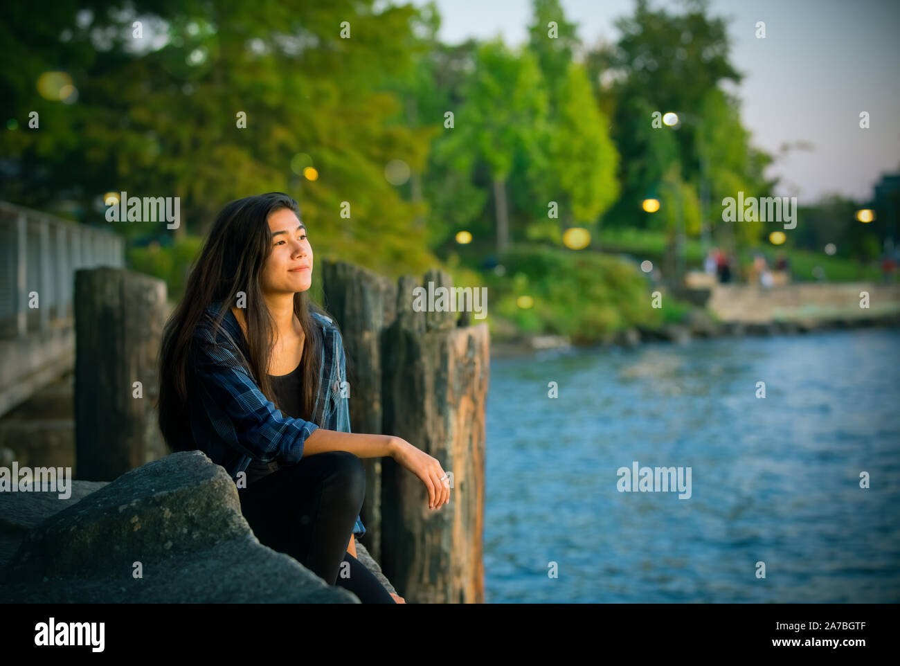 Biracial Asian Caucasian teen girl sitting by edge of lake on rocks looking out over lake at sunset Stock Photo