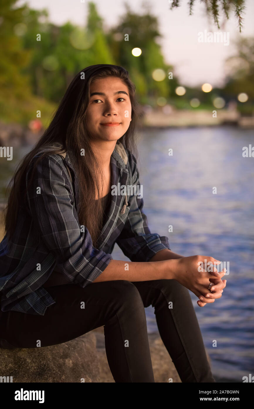 Biracial Asian Caucasian young teen girl sitting by edge of lake on ...