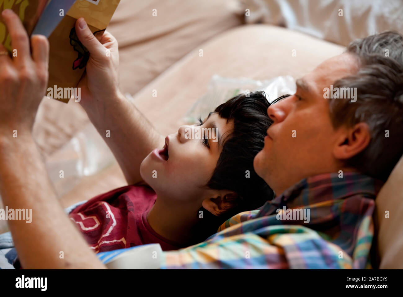Eight year old biracial boy reading book with Caucasian father while lying in bed Stock Photo