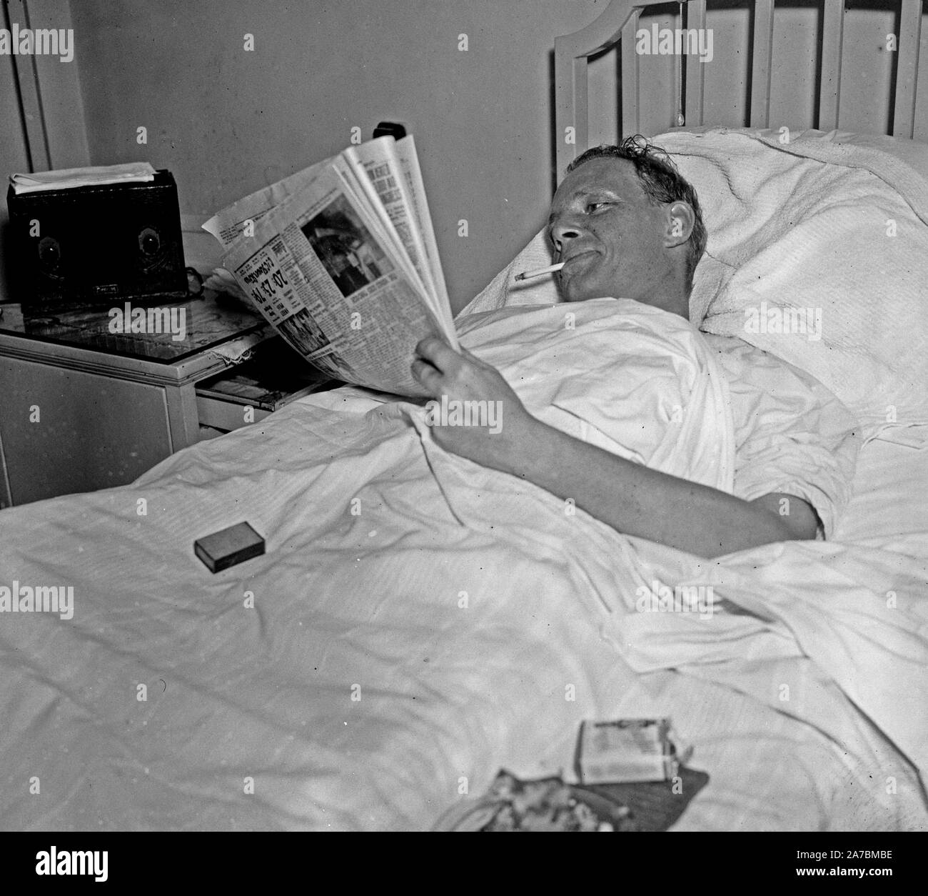 Man in hospital bed smoking a cigarette while reading a newspaper ca