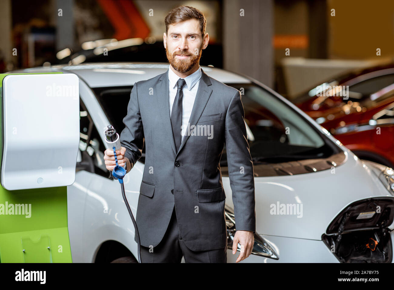 Portrait of a confident sales manager in the suit near the car charging station, selling electric cars in the showroom Stock Photo