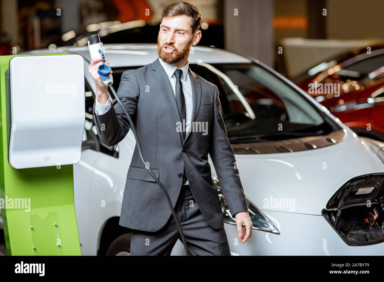 Funny portrait of a sales manager holding charging cable near the car charging station, selling electric cars in the showroom Stock Photo