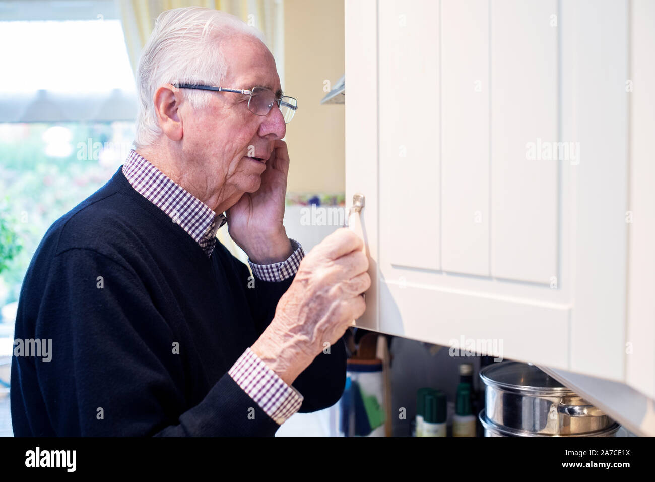 Forgetful Senior Man With Dementia Looking In Cupboard At Home Stock Photo