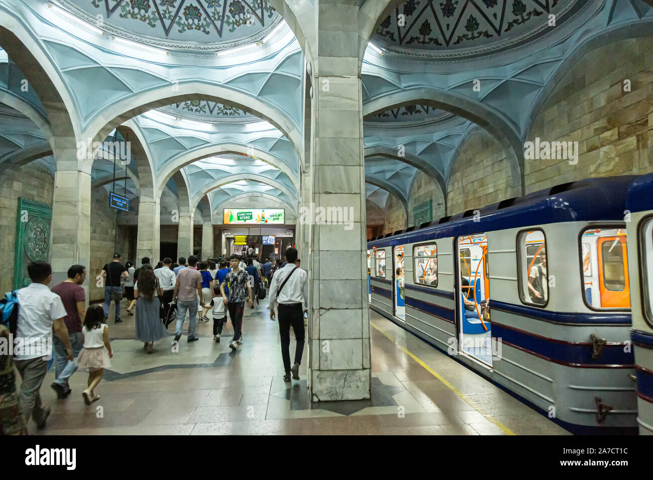 A number of commuters walking down the platform towards the exit of Alisher Navoi Metro Tashkent as a train sits at the station with its doors open. Stock Photo