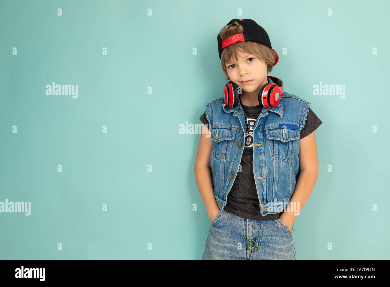A cool tenager boy in denim jaket and shorts, red earphones, black cap, stands in front of the camera and keeps his hands in pockets, isolated on blue Stock Photo