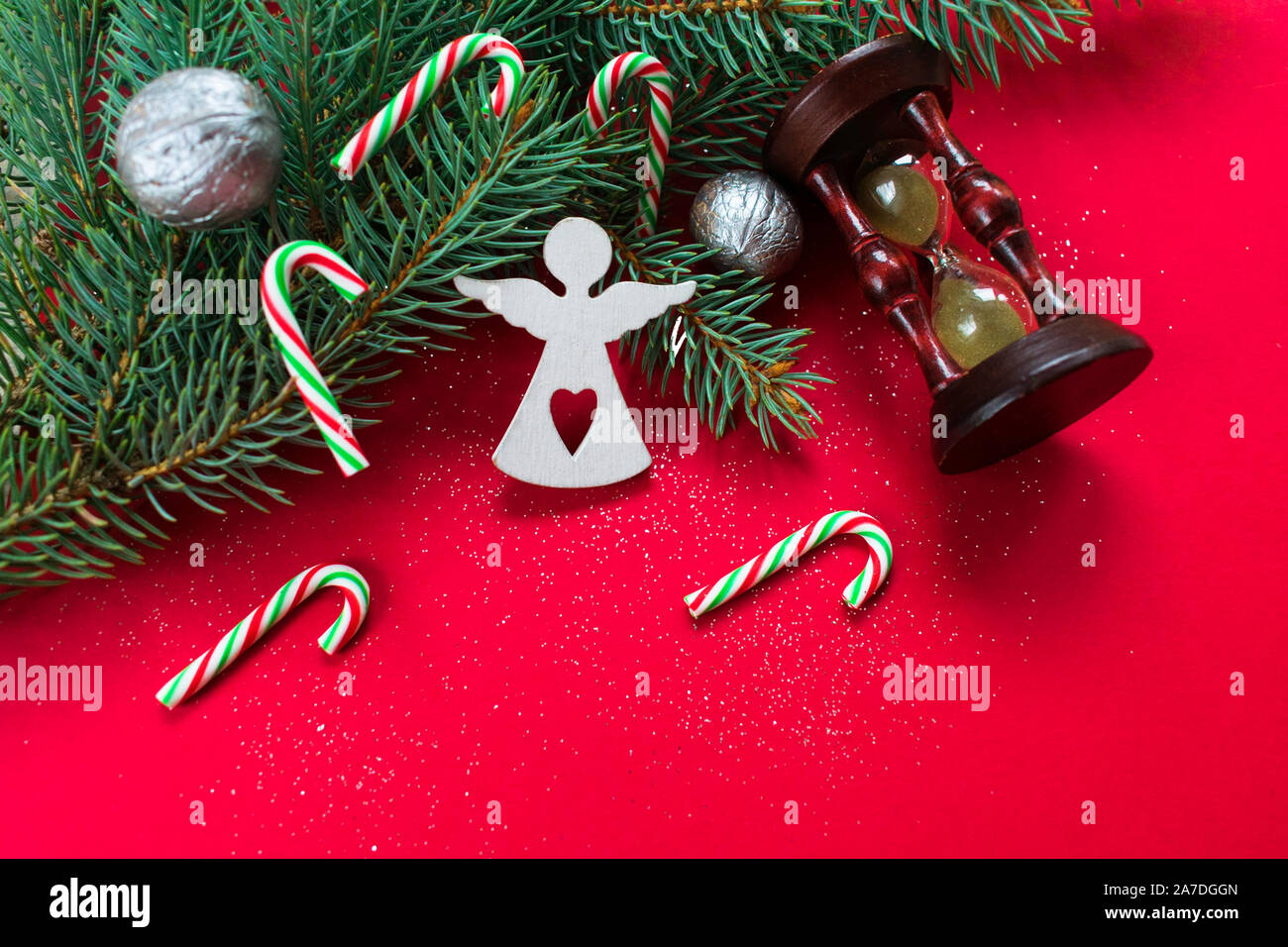 Christmas background on a red background with a Christmas branch of a yolka decorated with a figure of a white angel, sweets and sparkles. An hourglas Stock Photo