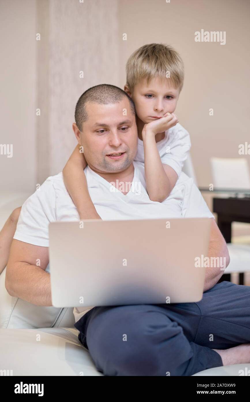 The son and his father are sitting on the couch, looking at a laptop, a happy family. Stock Photo