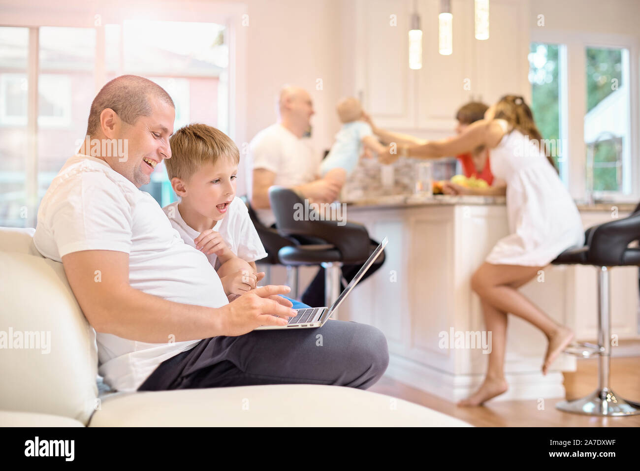 The son and his father are sitting on the couch, looking at the laptop, joyful emotions from what he saw, in the kitchen mom with friends and a small Stock Photo