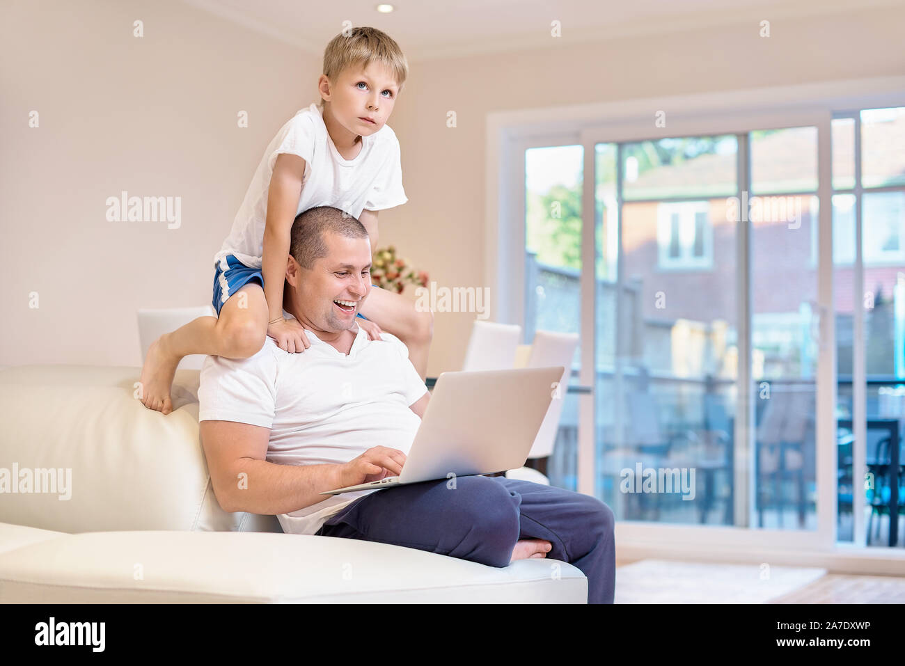 father sits on the couch, looks at the laptop and laughs, the son climbed on the neck to dad, emotions of joy from what he saw, happy family. Stock Photo
