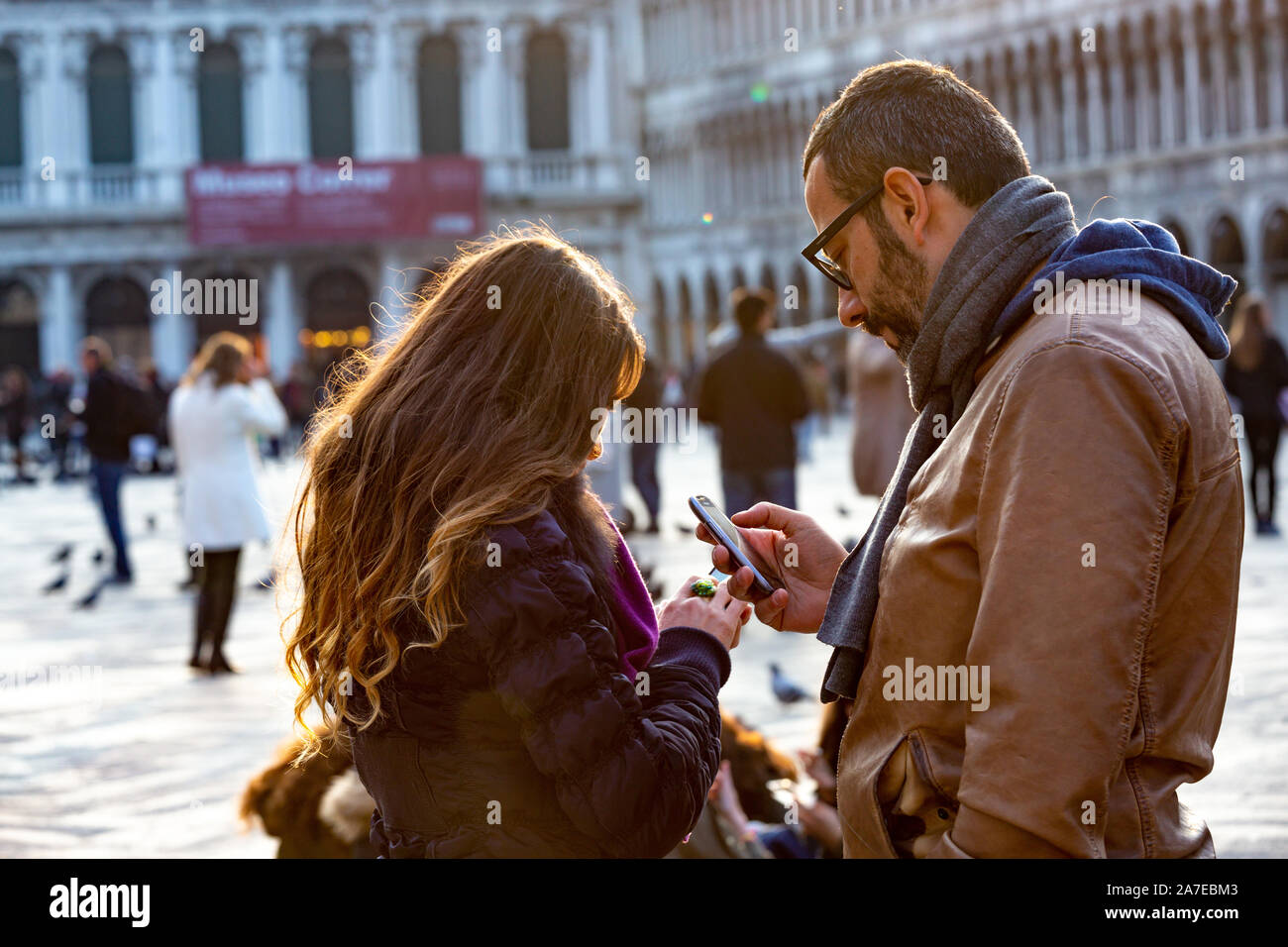 Venice, Italy - March 20, 2015:Side view of adult man and young woman browsing smartphones while standing in middle of Piazza San Marco during family Stock Photo