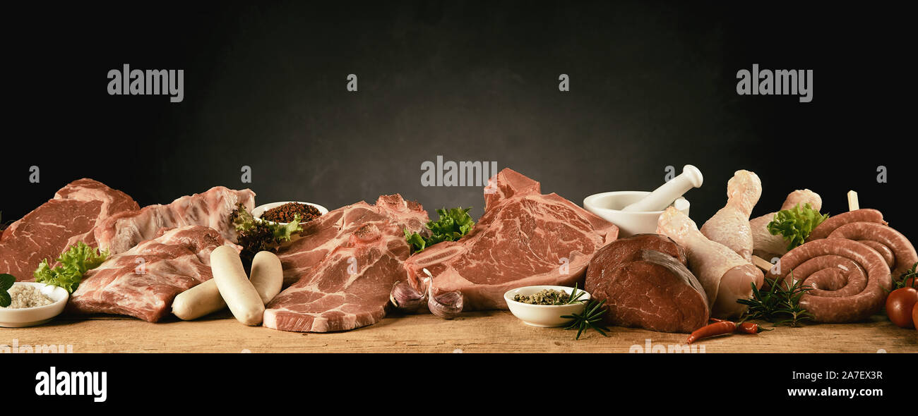 Raw assorted cuts of beef, pork, chicken and sausages for a barbecue displayed with fresh herbs and spices in a long row on a kitchen table in a panor Stock Photo
