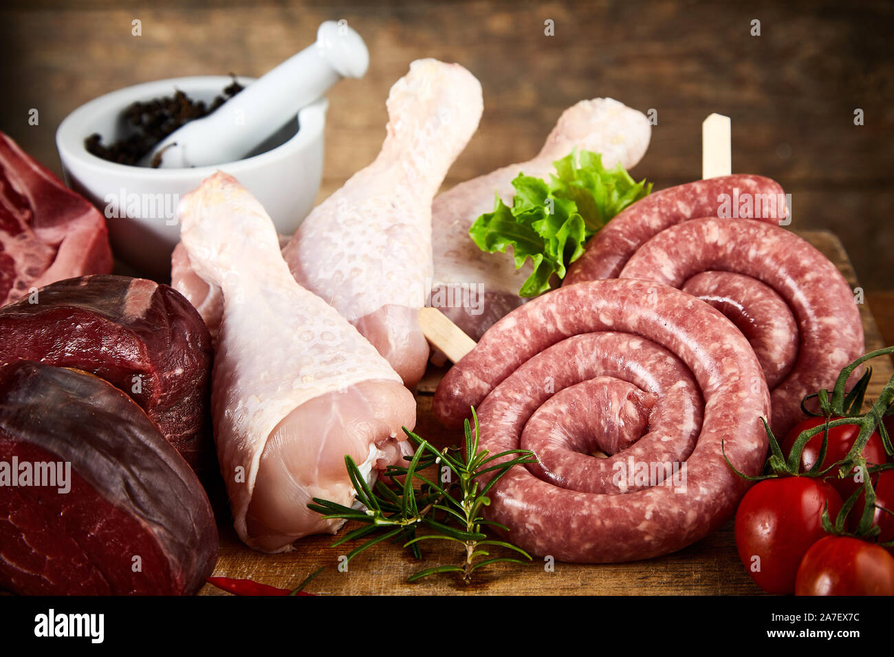 Rolled kringelwurst with raw chicken legs and beef steak displayed on a wooden kitchen table with fresh herbs and tomatoes in a close up view suitable Stock Photo