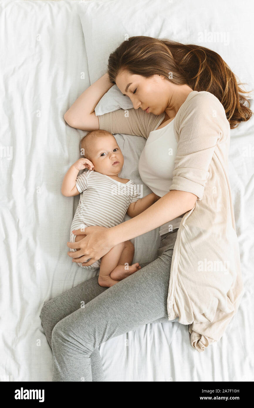Exhausted mother sleeping and embracing her awakened newborn baby Stock
