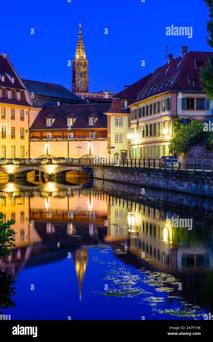 The steeple of Notre-Dame cathedral in Strasbourg, France, protrudes above the Petite France quarter at night and reflects in the water of the canal. Stock Photo