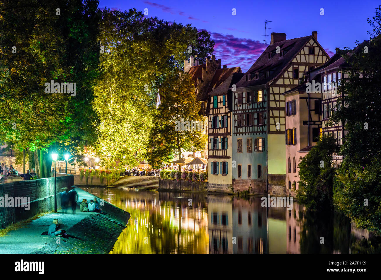 Young people enjoy the canal at night in the Petite France quarter in Strasbourg, France, opposite the half-timbered houses reflecting in the water. Stock Photo