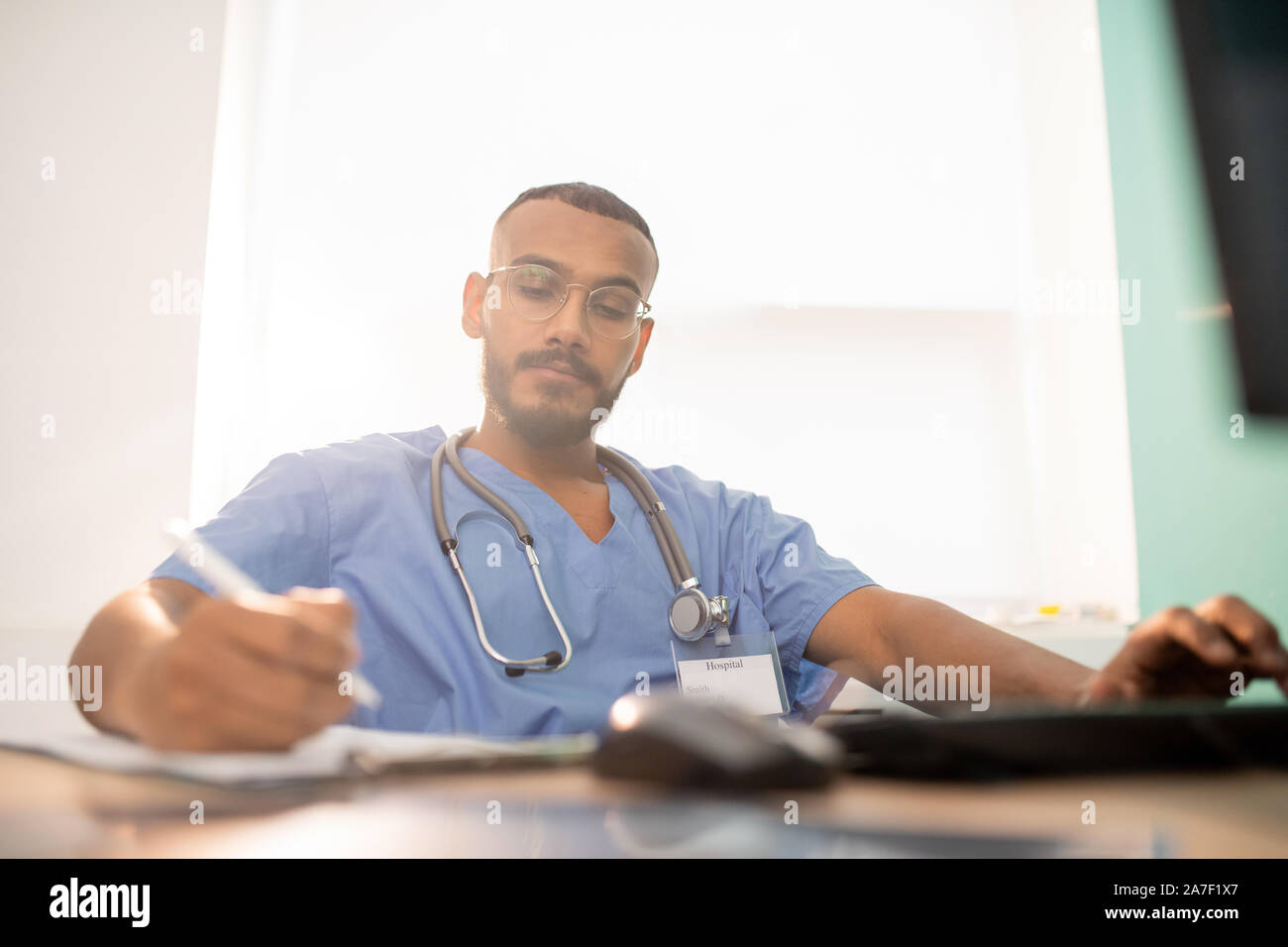 Bearded serious doctor or intern in uniform making working notes by table Stock Photo