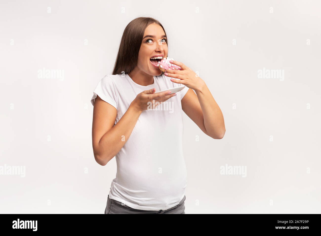 Excited Pregnant Lady Eating Donut Standing Over White Studio