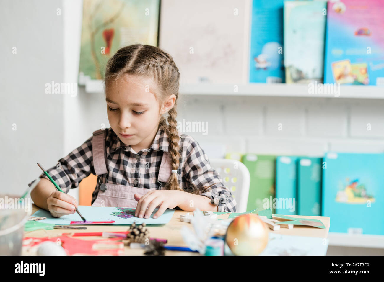 Creative schoolgirl with paintbrush sitting by desk and drawing xmas picture Stock Photo
