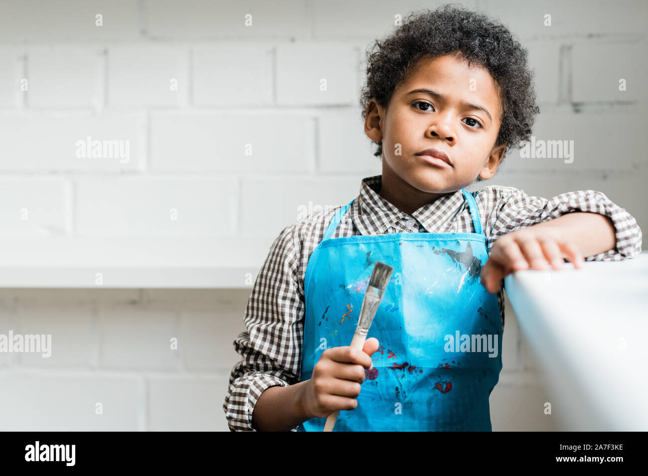 Serious African schoolboy in blue apron holding paintbrush in hand Stock Photo