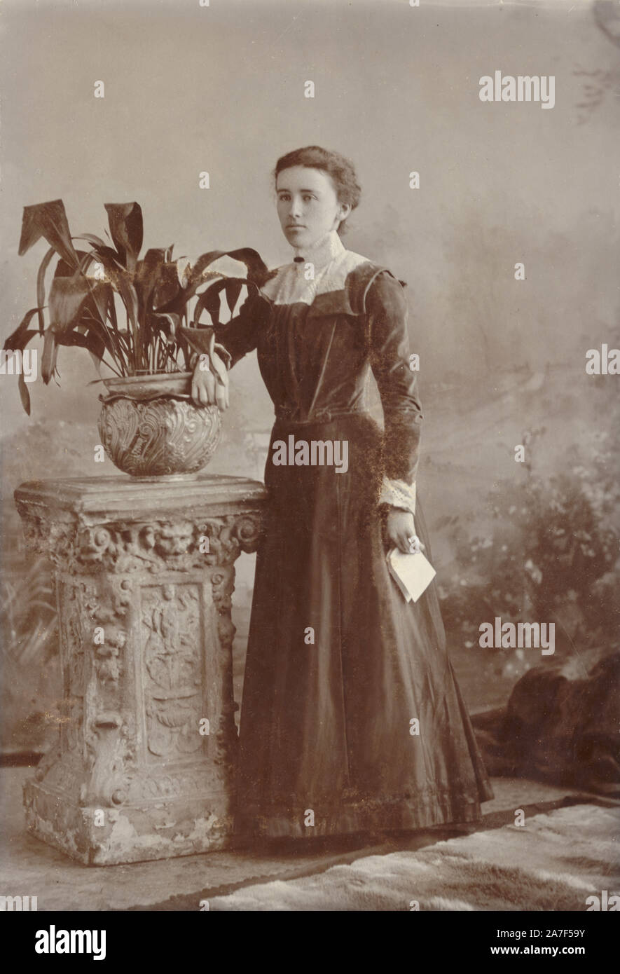 Late Victorian or early Edwardian cabinet photo card portrait of young Edwardian woman holding a letter and wearing a sombre, dark dress, posing next to potted plant - an aspidistra which were popular plants at this time. circa 1901, Truro, Cornwall, U.K. Stock Photo