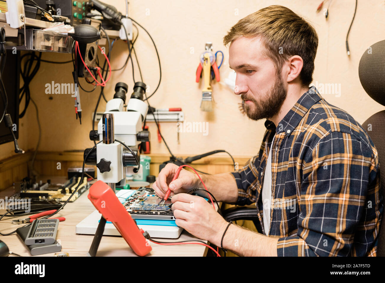 Young bearded mechanic with two soldering-irons sitting by workplace in workshop Stock Photo