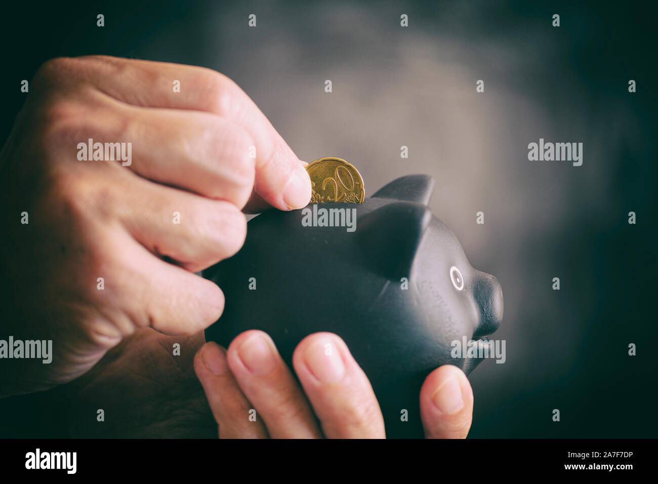 Person putting 20 euro cent coin into black piggy bank against black background. Close up. Stock Photo