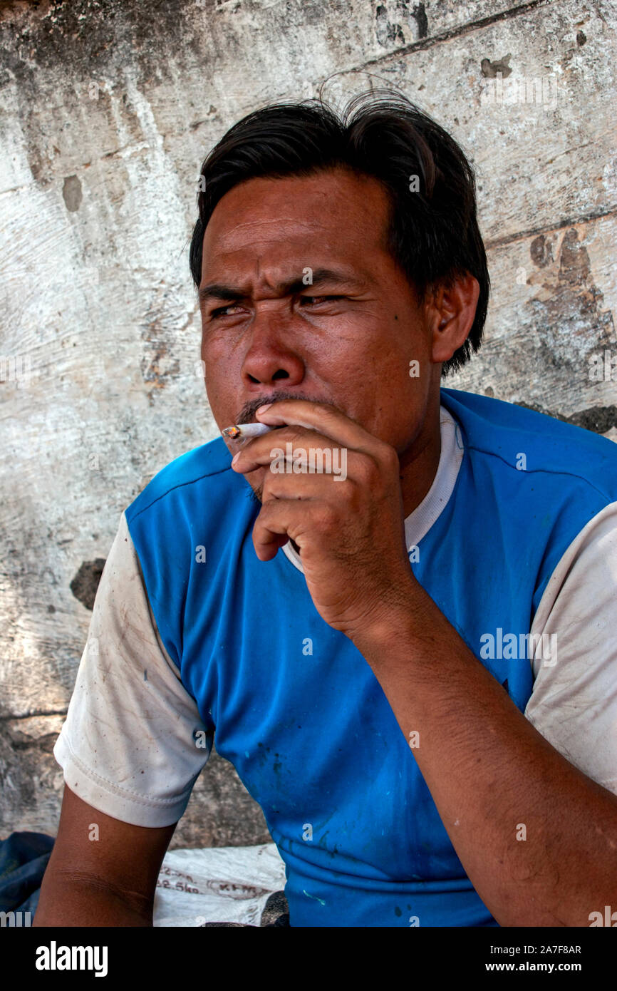 A young Thai man is smoking a cigarette while relaxing on a city street in Nong Khai, Northern Thailand. Stock Photo