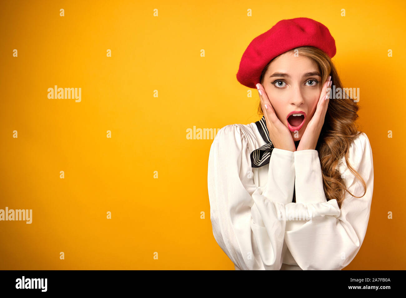 A girl with red lipstick in a white blouse and a red beret is standing opening her mouth in surprise, presses palms to her face Stock Photo