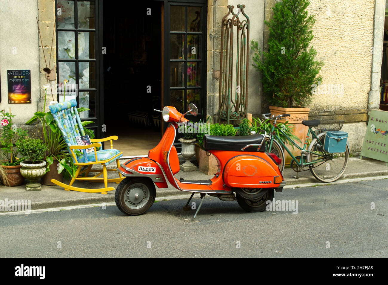 Orange scooter parked outside shop in Monpazier, France with old bicycle and chair Stock Photo