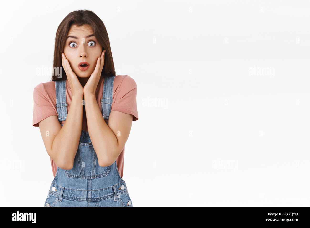 Shocked, speechless concerned young pity girl in overalls, t-shirt, holding hands on cheeks stare camera with popped eyes, gasping amazed, heard Stock Photo