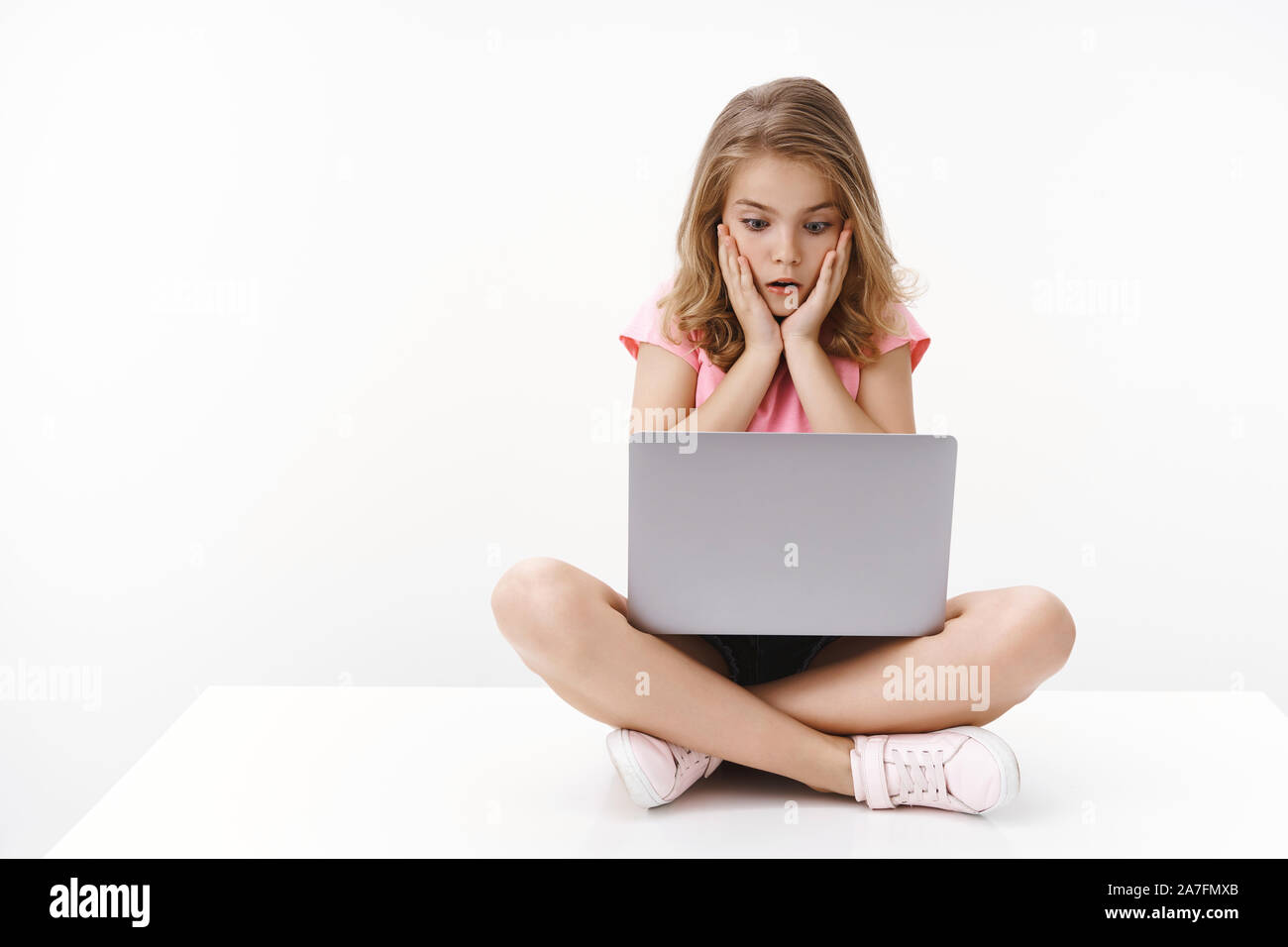 Shocked young blond girl child sitting crossed legs, watching scary video internet, hold laptop, gasping shook stare computer screen, touch cheeks Stock Photo